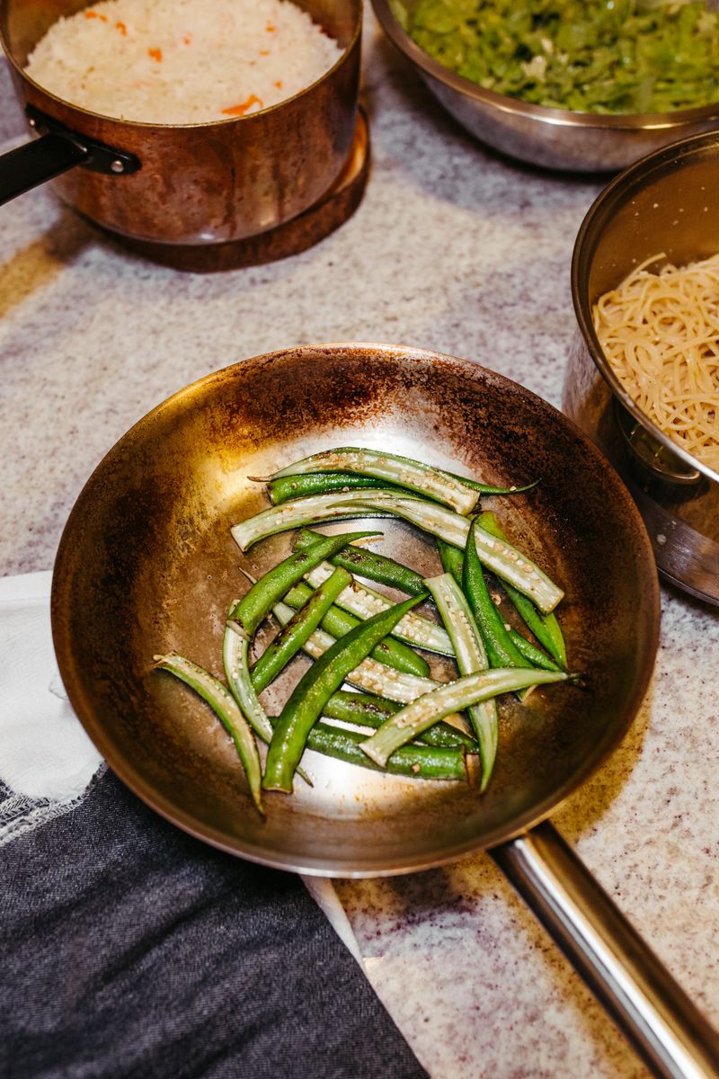 Fresh okra sizzling in a copper pan, accompanied by rice and pasta ingredients